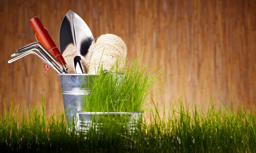 A close-up view of a gardening scene with two small metal containers placed on a lush green lawn area. One container holds various hand gardening tools, including a trowel, pruners, and a rake, with a ball of gardening twine resting on top. The background features a warm-toned wooden surface, possibly fencing or a garden shed, providing a natural backdrop. The scene emphasizes preparedness and care in outdoor garden maintenance, typical of professional gardening services offered in Southwark, with the vibrant green grass contrasting against the metallic tools and earthy tones of the background.