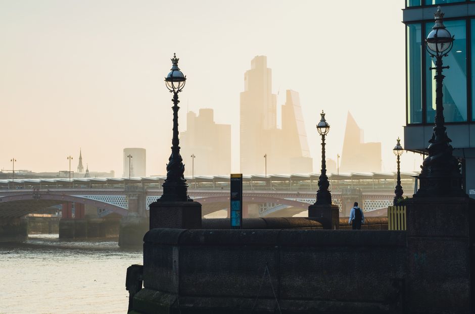 A riverside scene featuring a paved promenade with ornate black cast-iron street lamps lining the edge of the water. In the background, a modern urban skyline with tall glass skyscrapers is partially