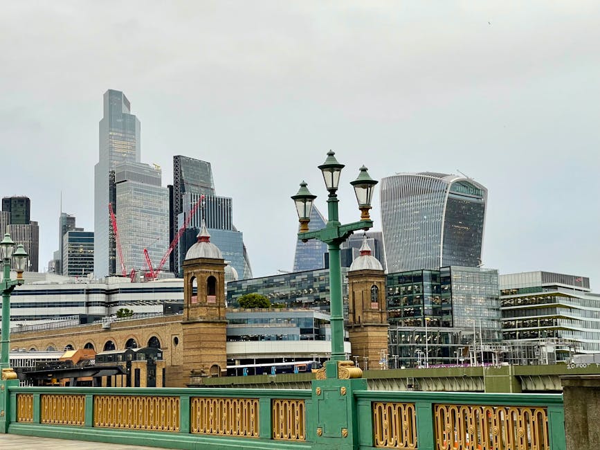 A riverside scene featuring a paved promenade with ornate black cast-iron street lamps lining the edge of the water. In the background, a modern urban skyline with tall glass skyscrapers is partially